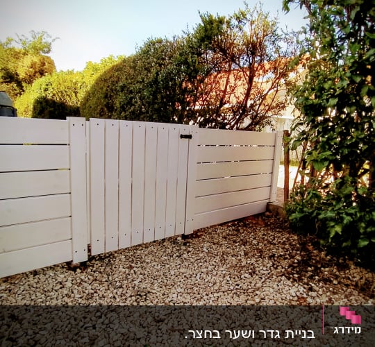 גדר עץ לבנה על חצץ ליד צמחייה ירוקה